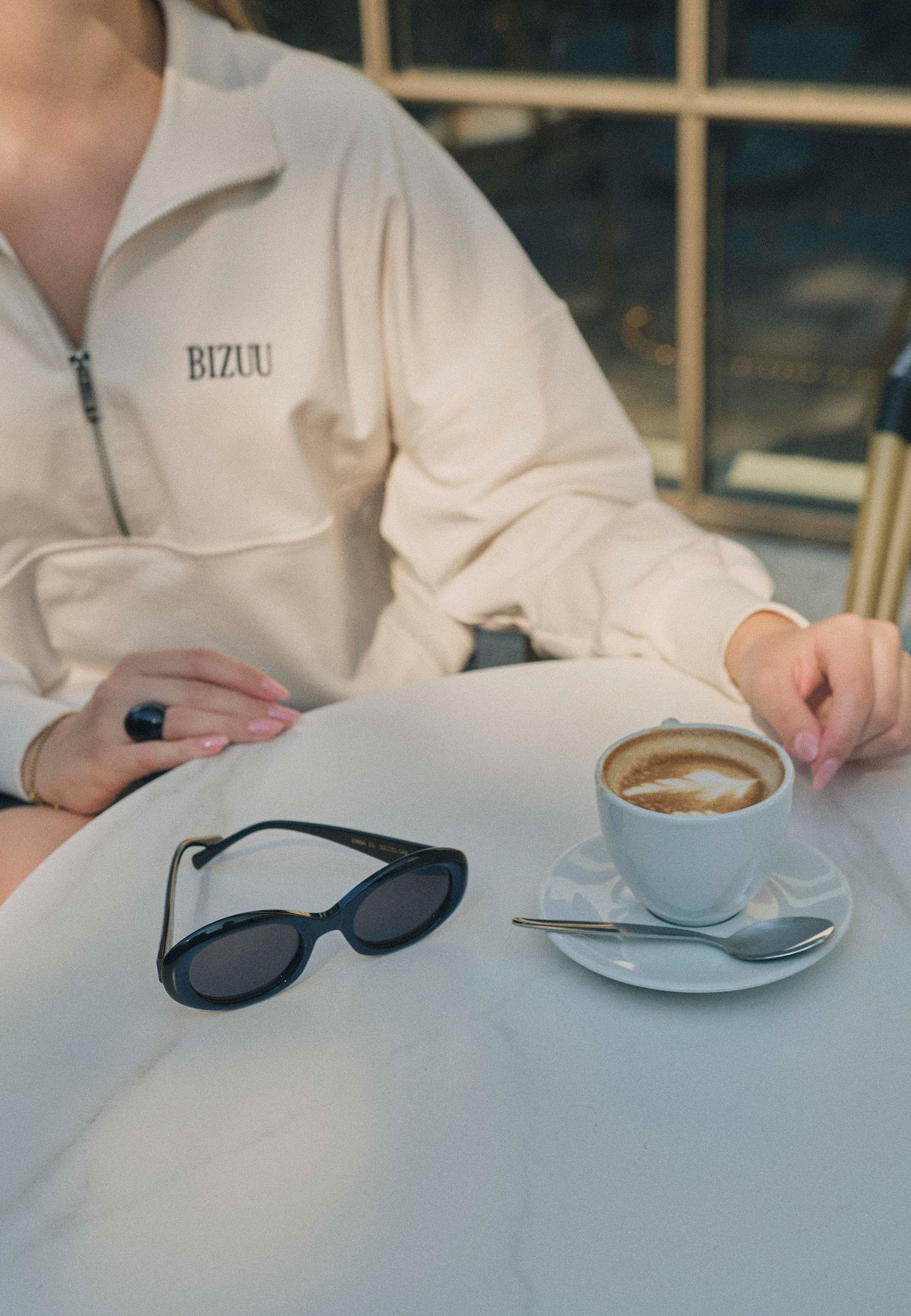 Zbliżenie na biały stolik z filiżanką cappuccino i odłożonymi czarnymi okularami BIZUU ENNA. Na pierwszym planie ręce kobiety w jasnej bluzie z logo BIZUU i czarnym pierścionkiem.