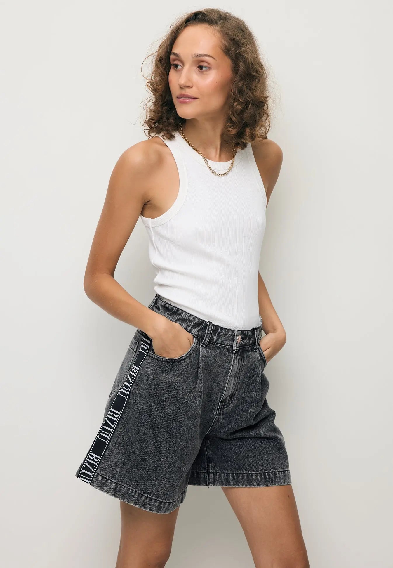 Modelka stojąca tyłem w białym topie ALEXIN z logo na plecach oraz jeansowych szortach MIREYA.