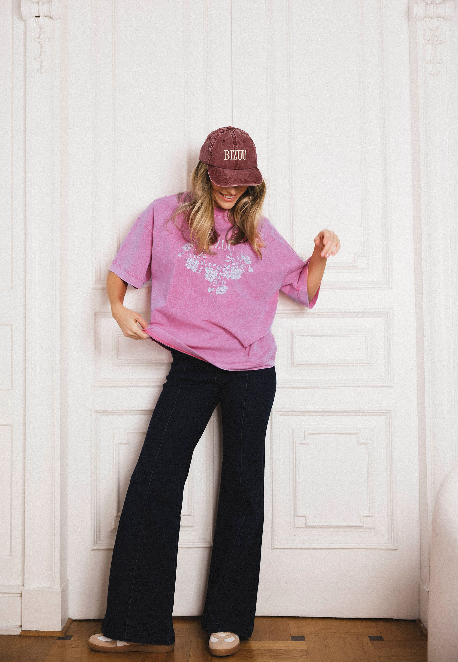 Modelka na tle białych drzwi w różowej koszulce oversize PAPLYS i jeansach flare BELINNA.