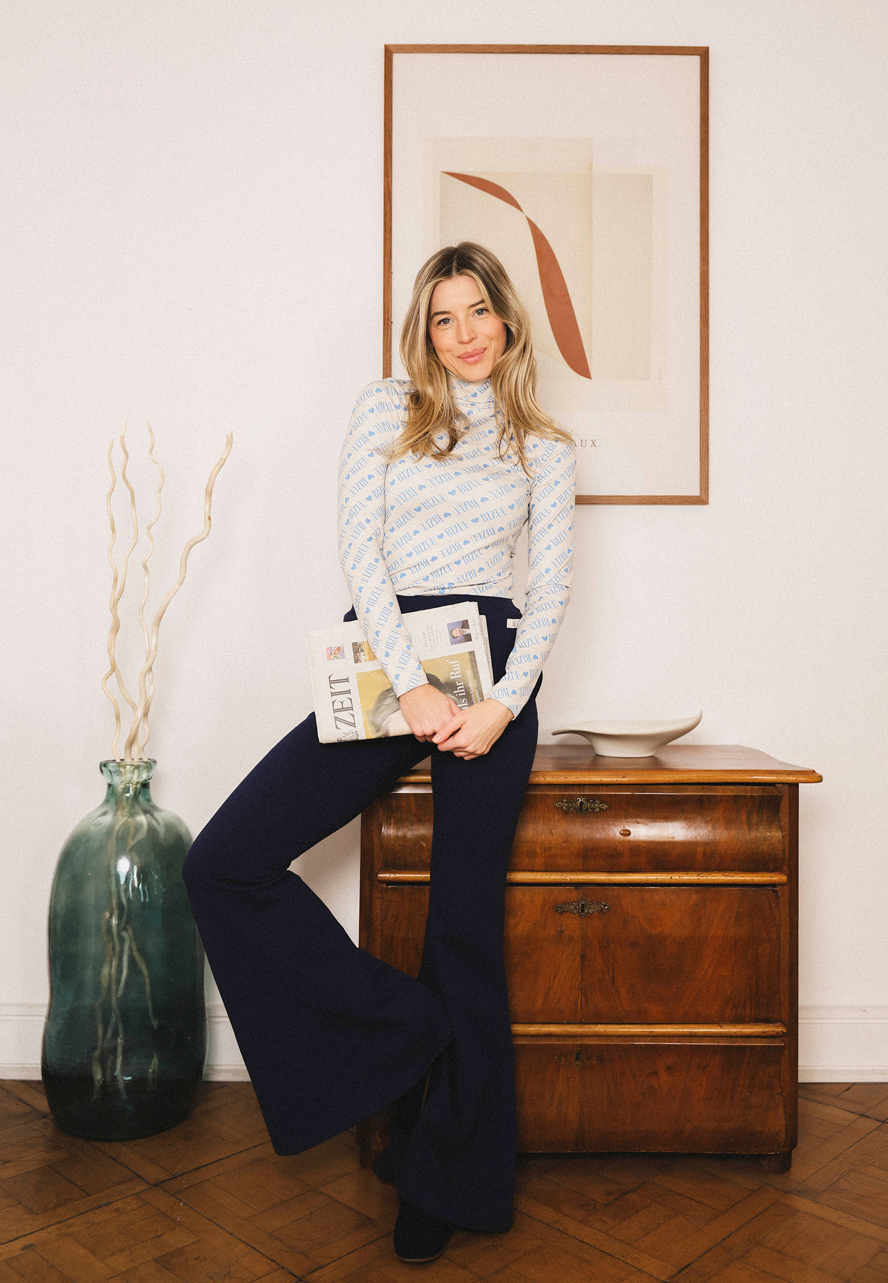 Modelka oparta o komodę ubrana w biały, obcisły golf SOPHIE i granatowe spodnie dresowe TEIA.