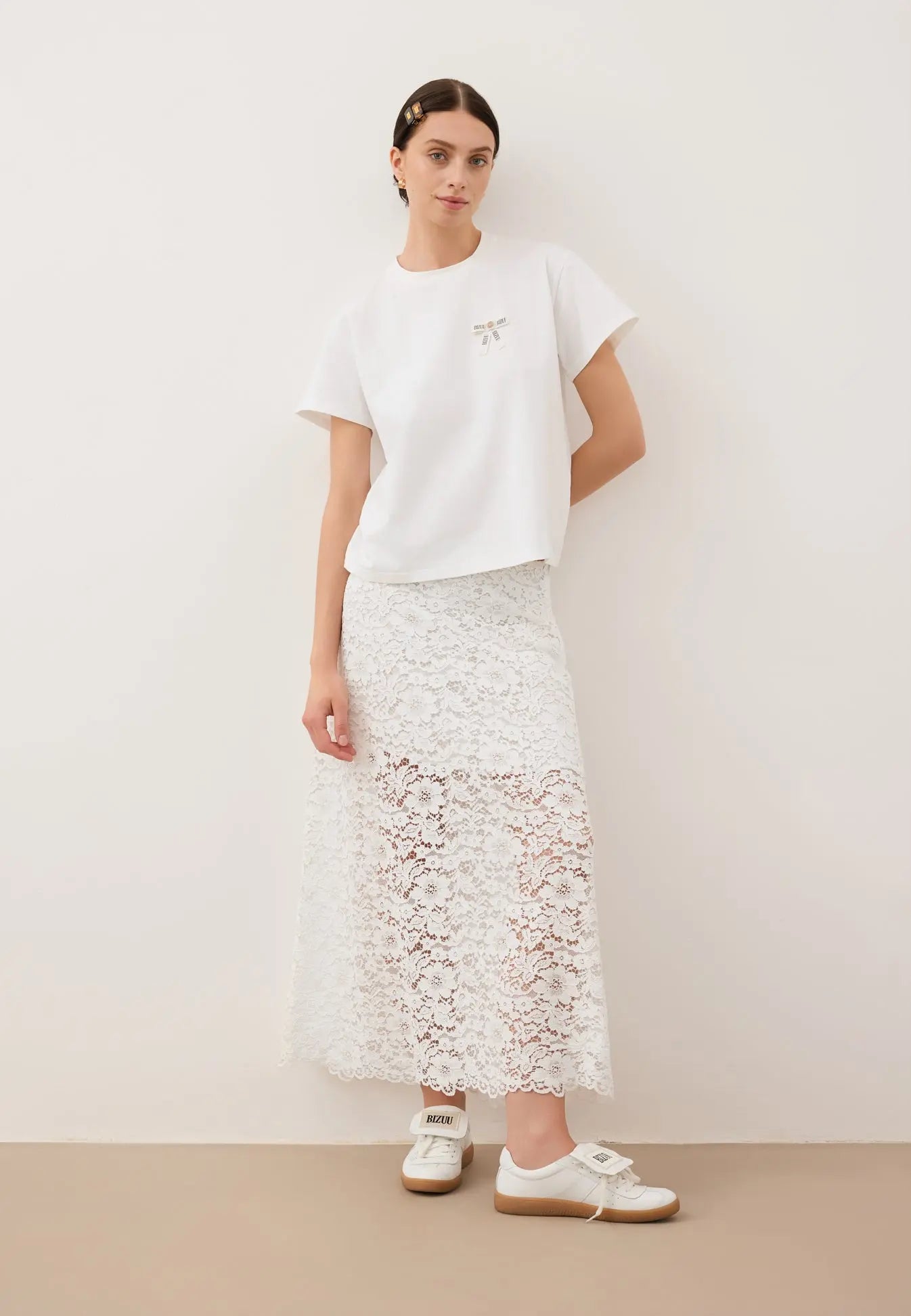 Modelka oparta o białą ścianę, ubrana w biały T-shirt z logowaną kokardką TISHALLE i białą, koronkową spódniczkę MILKY.